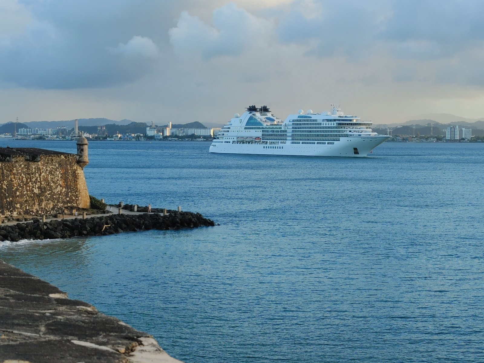 A cruise ship on calm water at dusk near a harbour, with a stone fort and the port town behind it.